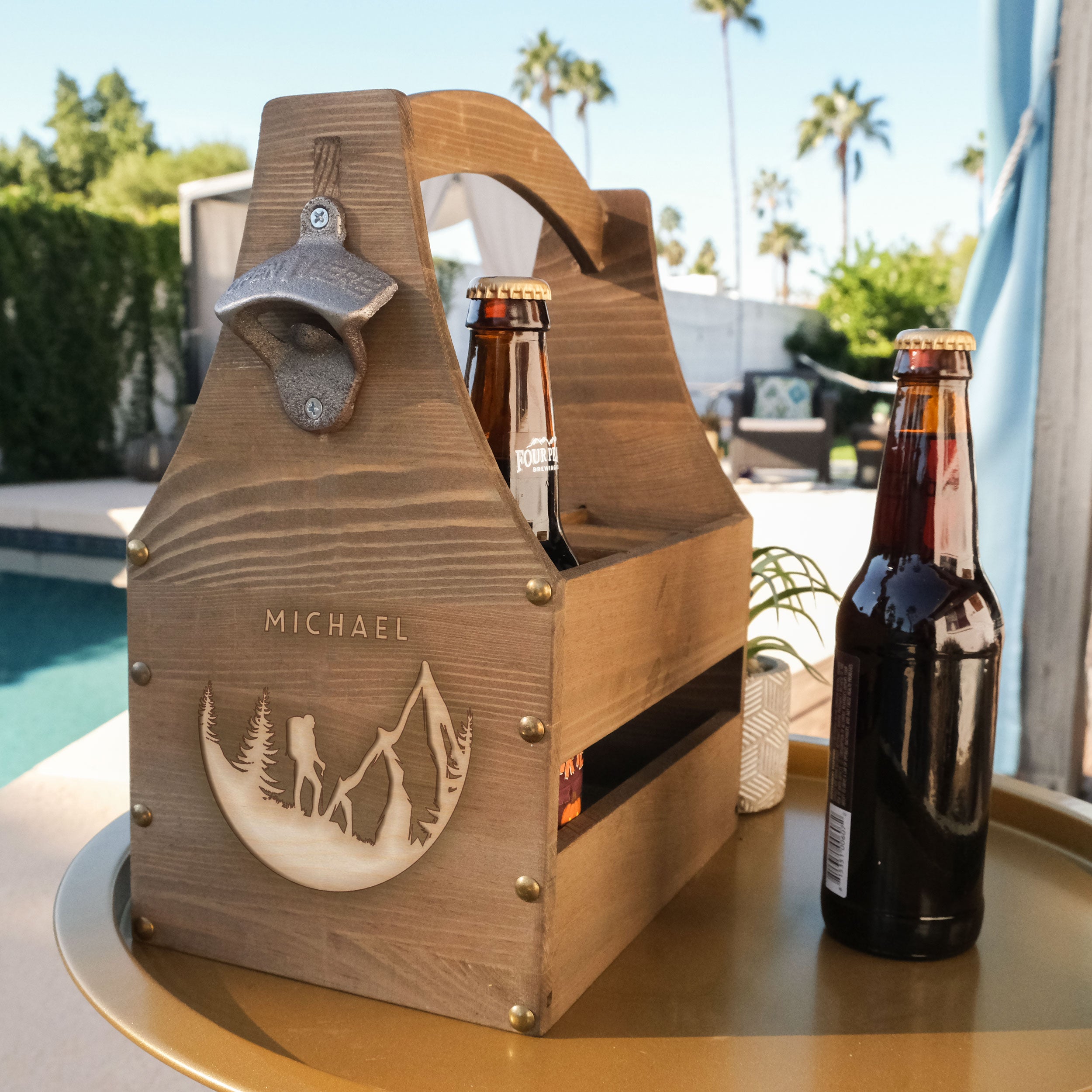 Wood beer caddy on a table. The beer caddy has a design engraved on one side. The design has "MICHAEL" in print font centered at the top. Below is a circular image of a hiker and mountains and forrest trees. The design is centered towards the bottom.