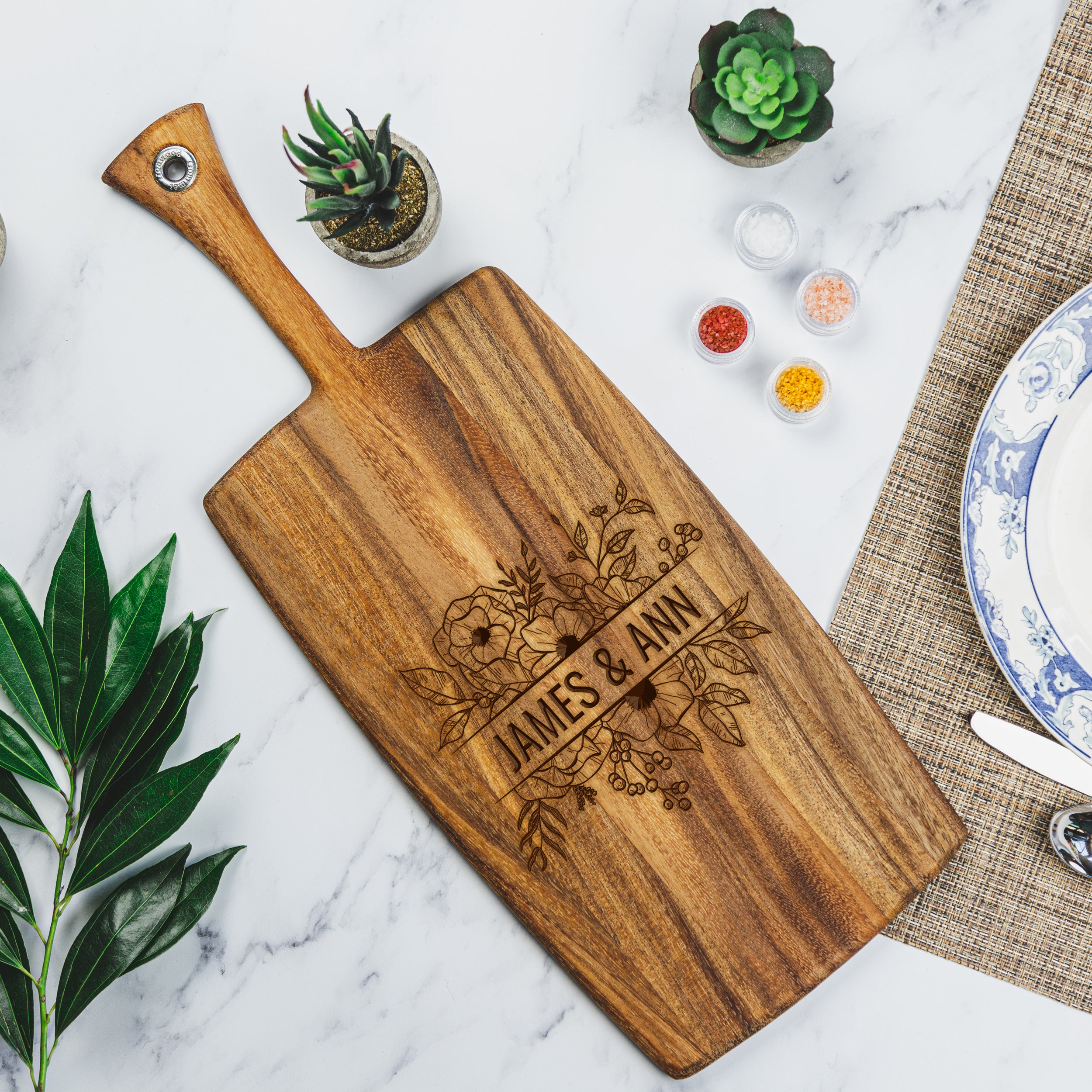 Birds eye view of a charcuterie board on a marble table top. The charcuterie board is wood, and is a long rectangular board with a handle. The board is engraved with a floral design and the names "JAMES & ANN" in the middle.