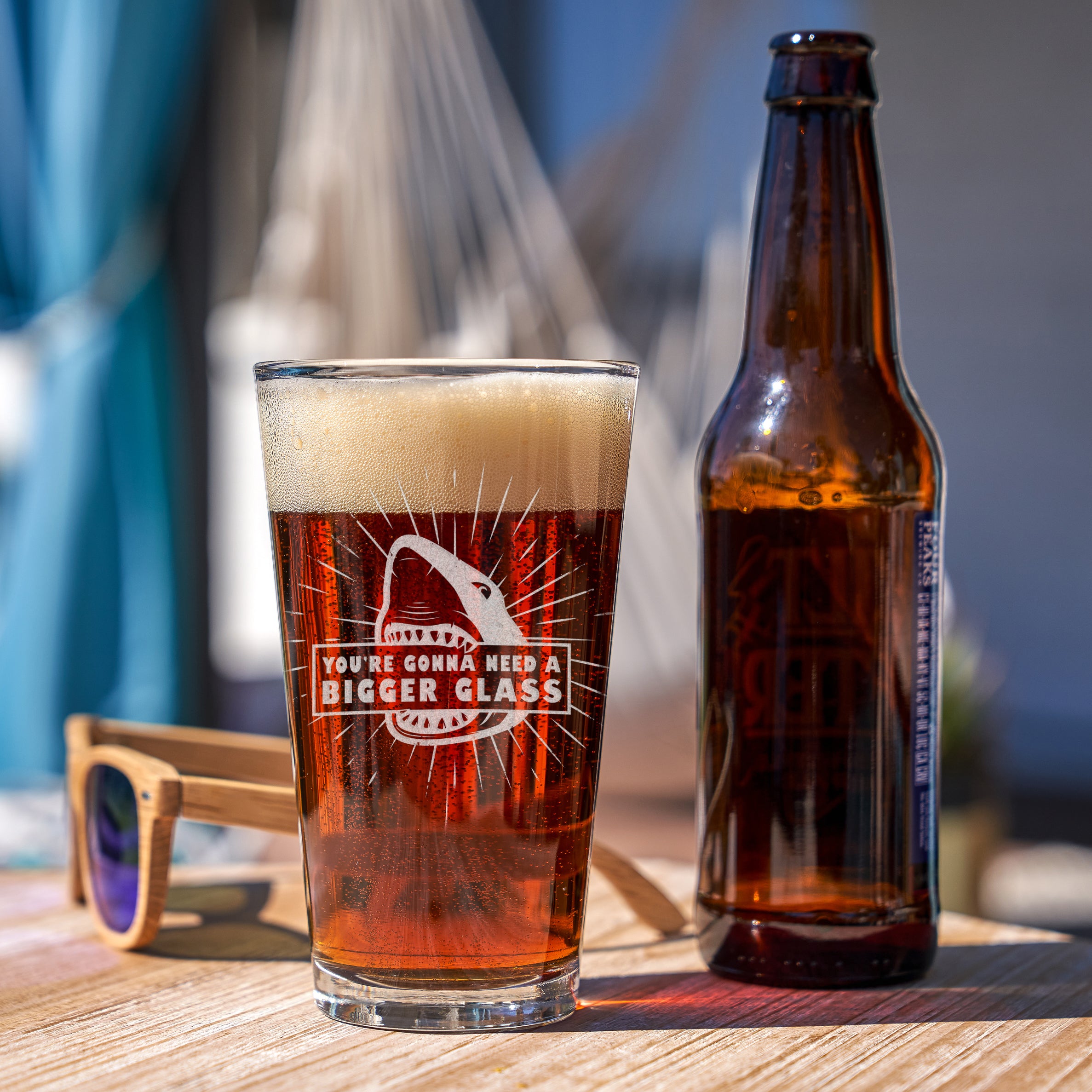 Pint glass on a table next to a beer bottle. The glass has a etched design on the front and in the center. The design is of a shark, with radial lines surrounding it. In the sharks mouth is a sign that says "YOU'RE GONNA NEED A BIGGER GLASS" in print font. The design is a reference to the movie JAWS.