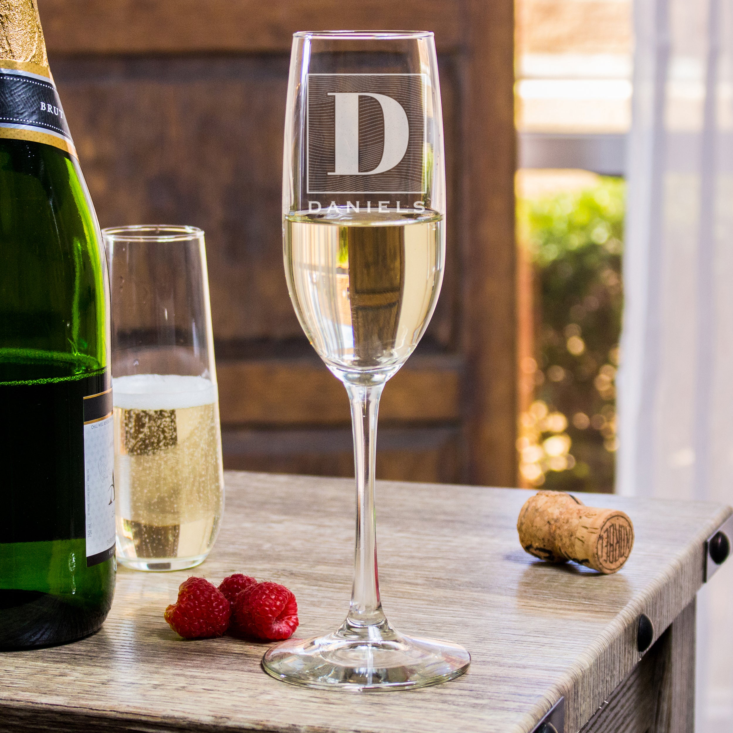 Champagne glass on a table. The glass has an etched design centered. The design is of a square with swirly horizontal lines, and in the middle is the letter "D" in caps. Below the square is the name "DANIELS" in all caps. 