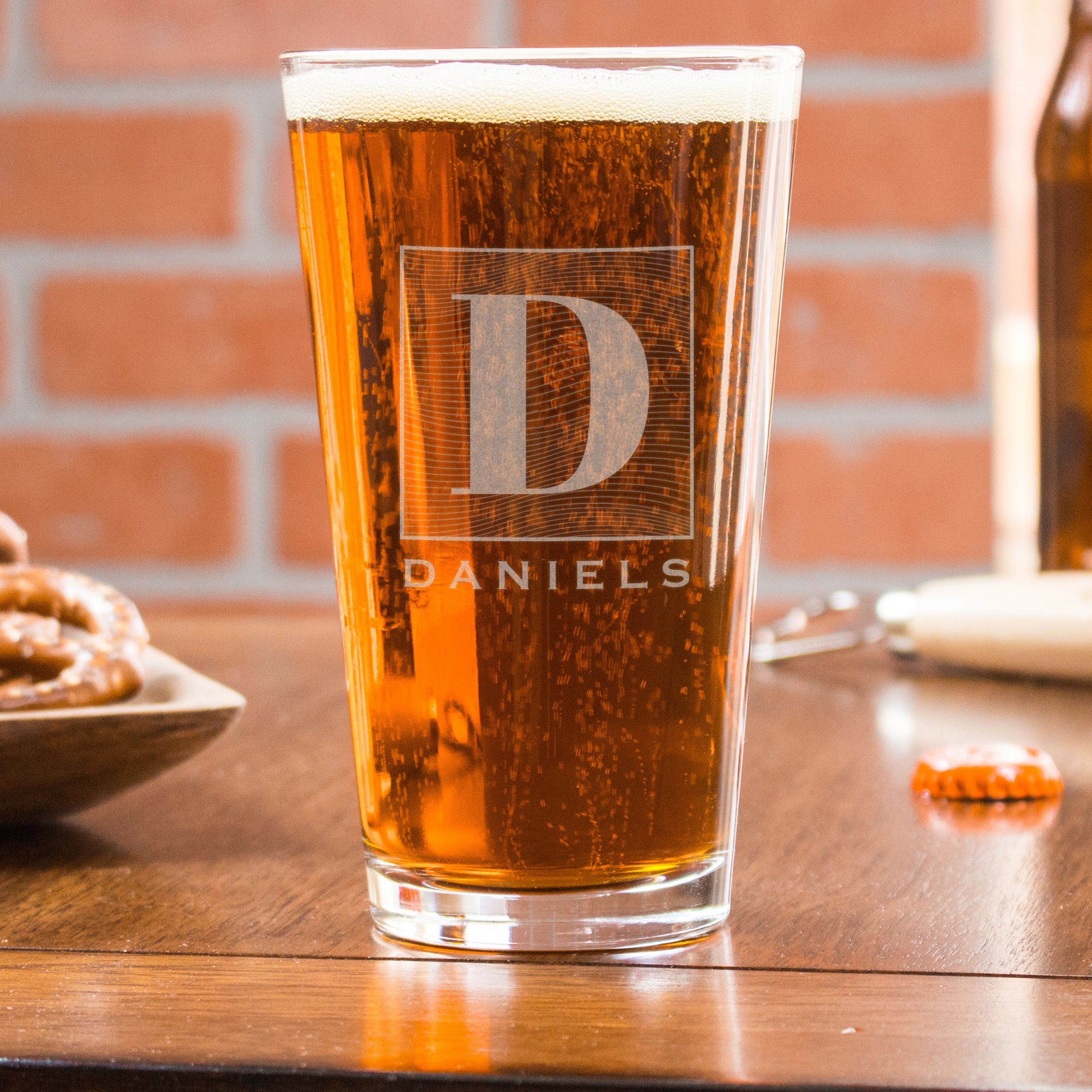 Pint glass on a table. The glass has an etched design centered. The design is of a square with swirly horizontal lines, and in the middle is the letter "D" in caps. Below the square is the name "DANIELS" in all caps. 