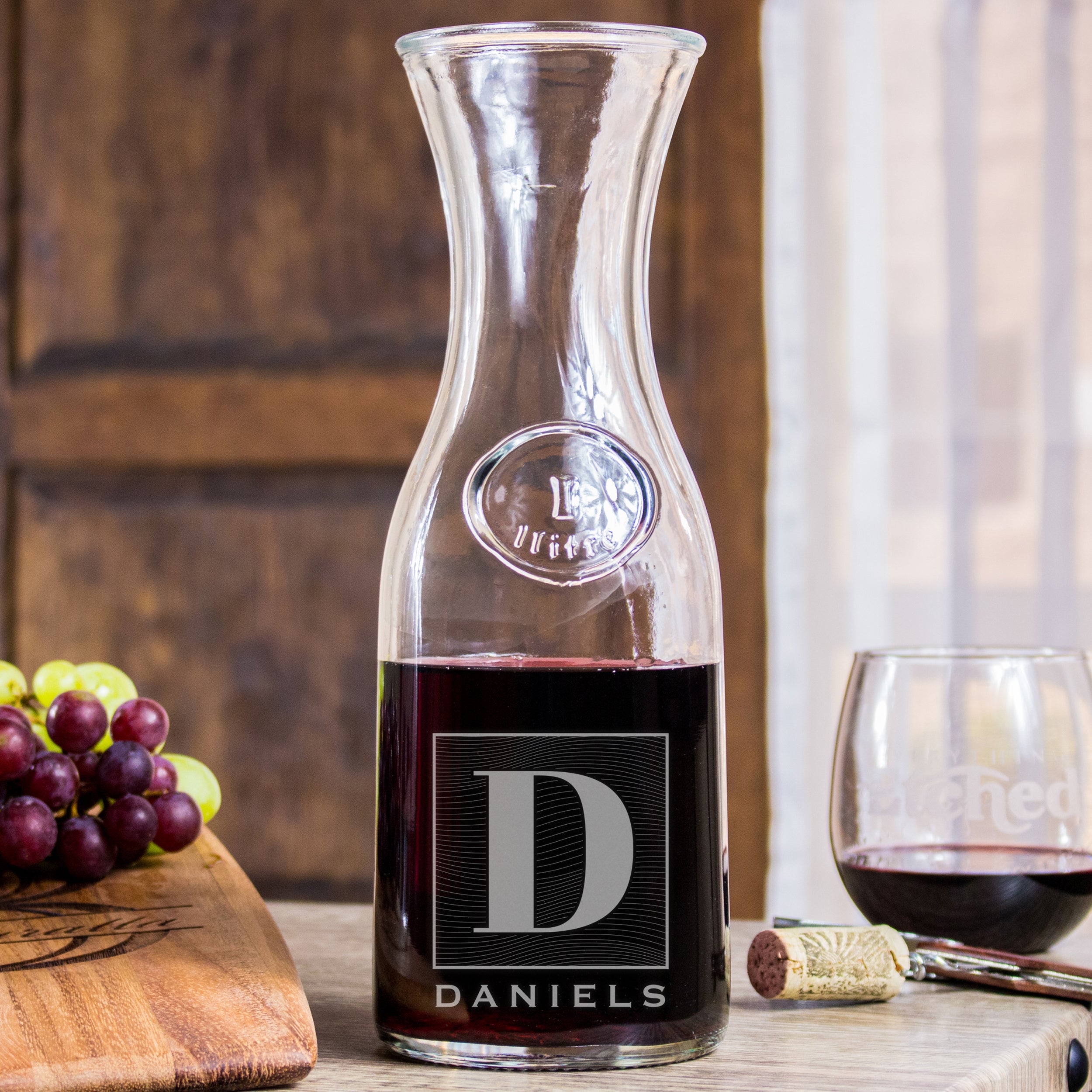 Glass wine decanter on a table. The glass has an etched design centered towards the bottom. The design is of a square with swirly horizontal lines, and in the middle is the letter "D" in caps. Below the square is the name "DANIELS" in all caps. 