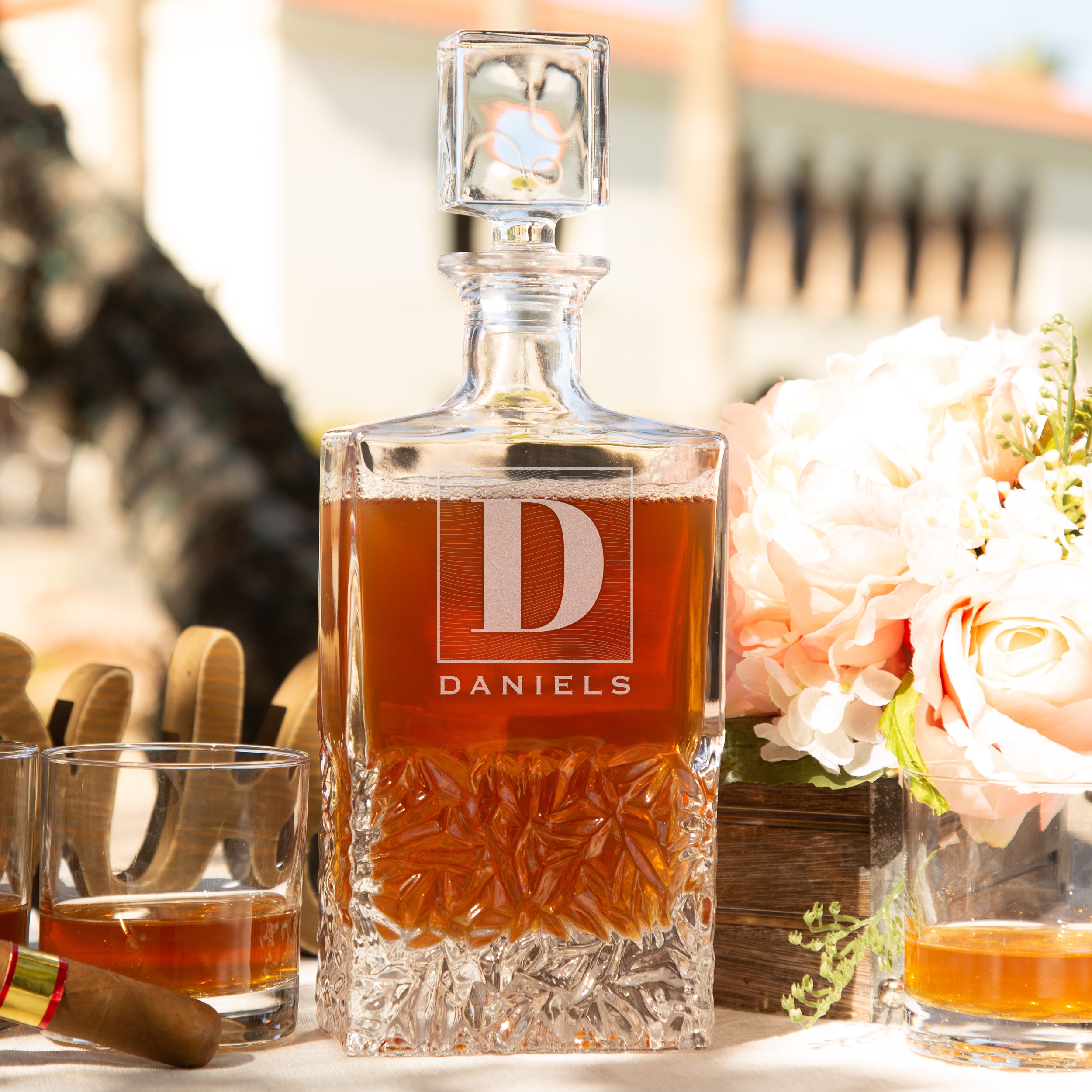 Glass whiskey decanter on a table. The glass has an etched design centered at the top. The design is of a square with swirly horizontal lines, and in the middle is the letter "D" in caps. Below the square is the name "DANIELS" in all caps. 