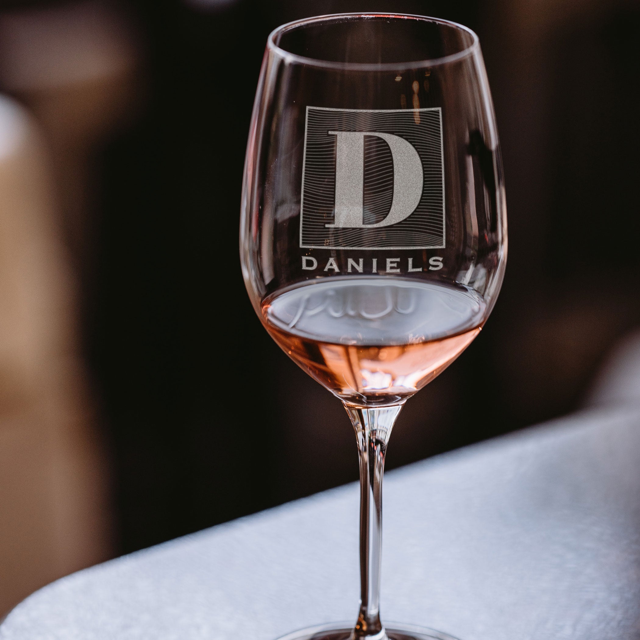 Stemmed wine glass on a table. The glass has an etched design centered. The design is of a square with swirly horizontal lines, and in the middle is the letter "D" in caps. Below the square is the name "DANIELS" in all caps. 