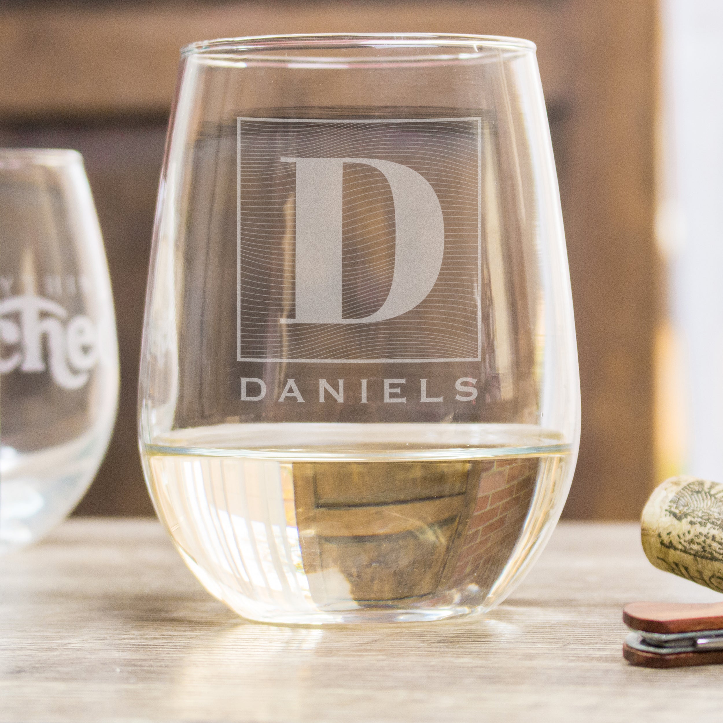 Stemless white wine glass on a table. The glass has an etched design centered. The design is of a square with swirly horizontal lines, and in the middle is the letter "D" in caps. Below the square is the name "DANIELS" in all caps. 