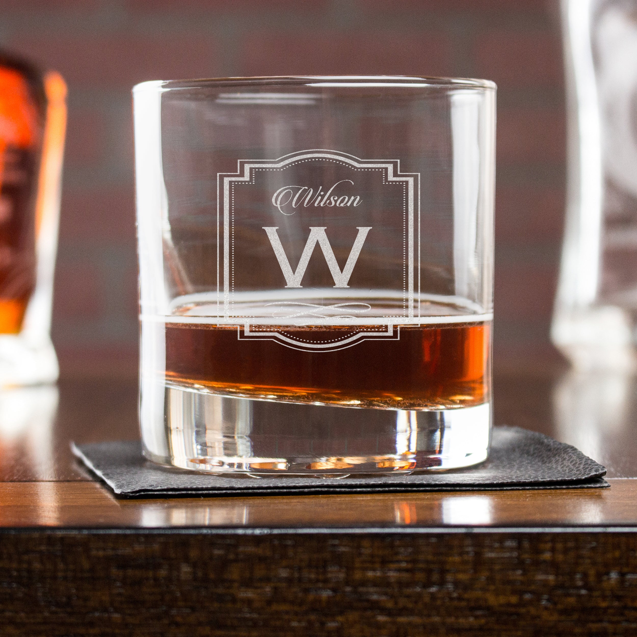 Etched whiskey glass on a table. The glass is etched with a design centered on the glass. The design is of "Wilson" is cursive, and below it is a capital "W" and underneath it is a decorative swirly line. Around the entire design is a decorative border of lines and dots. 