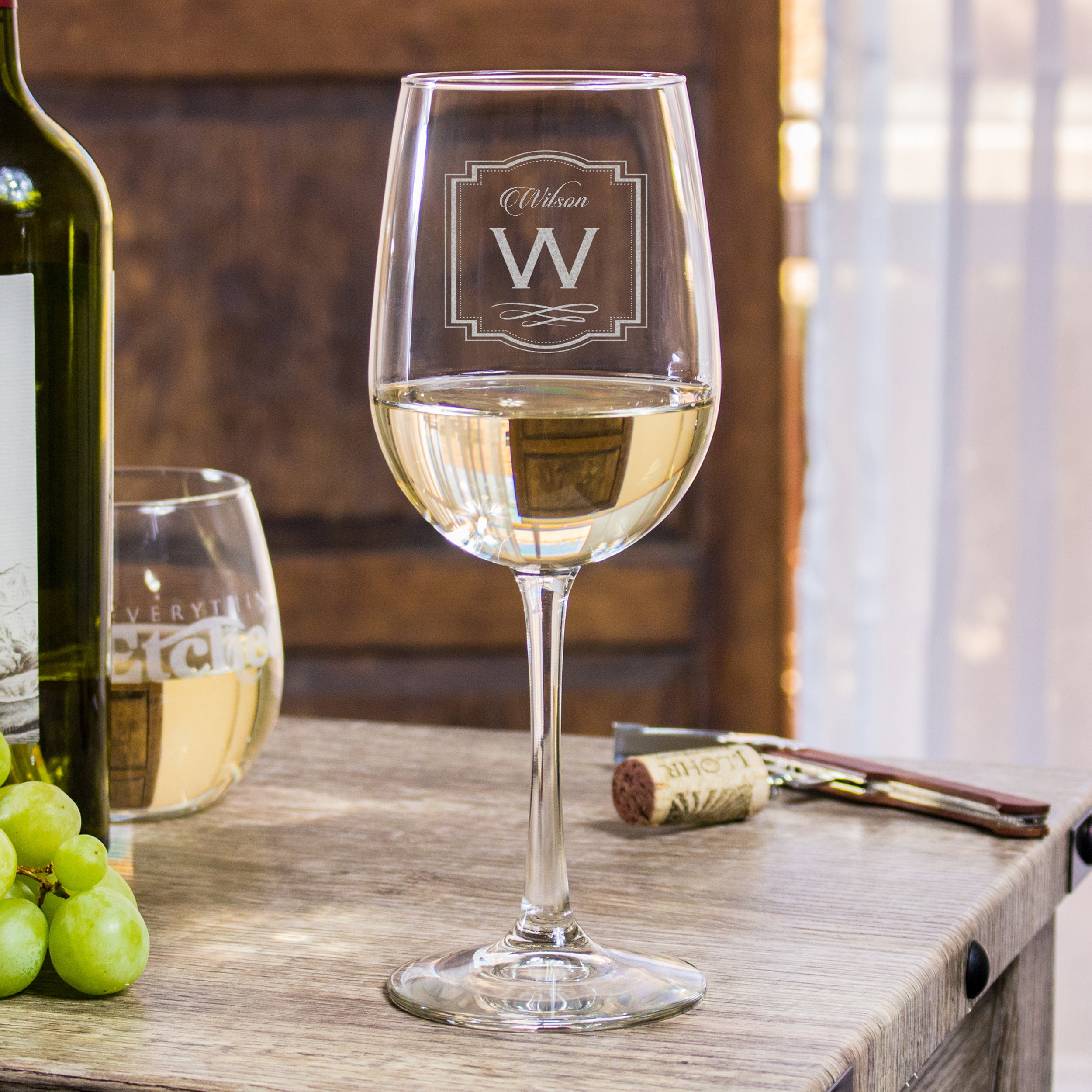 Stemmed white wine glass on a table. The glass is etched with a design centered on the glass. The design is of "Wilson" is cursive, and below it is a capital "W" and underneath it is a decorative swirly line. Around the entire design is a decorative border of lines and dots. 