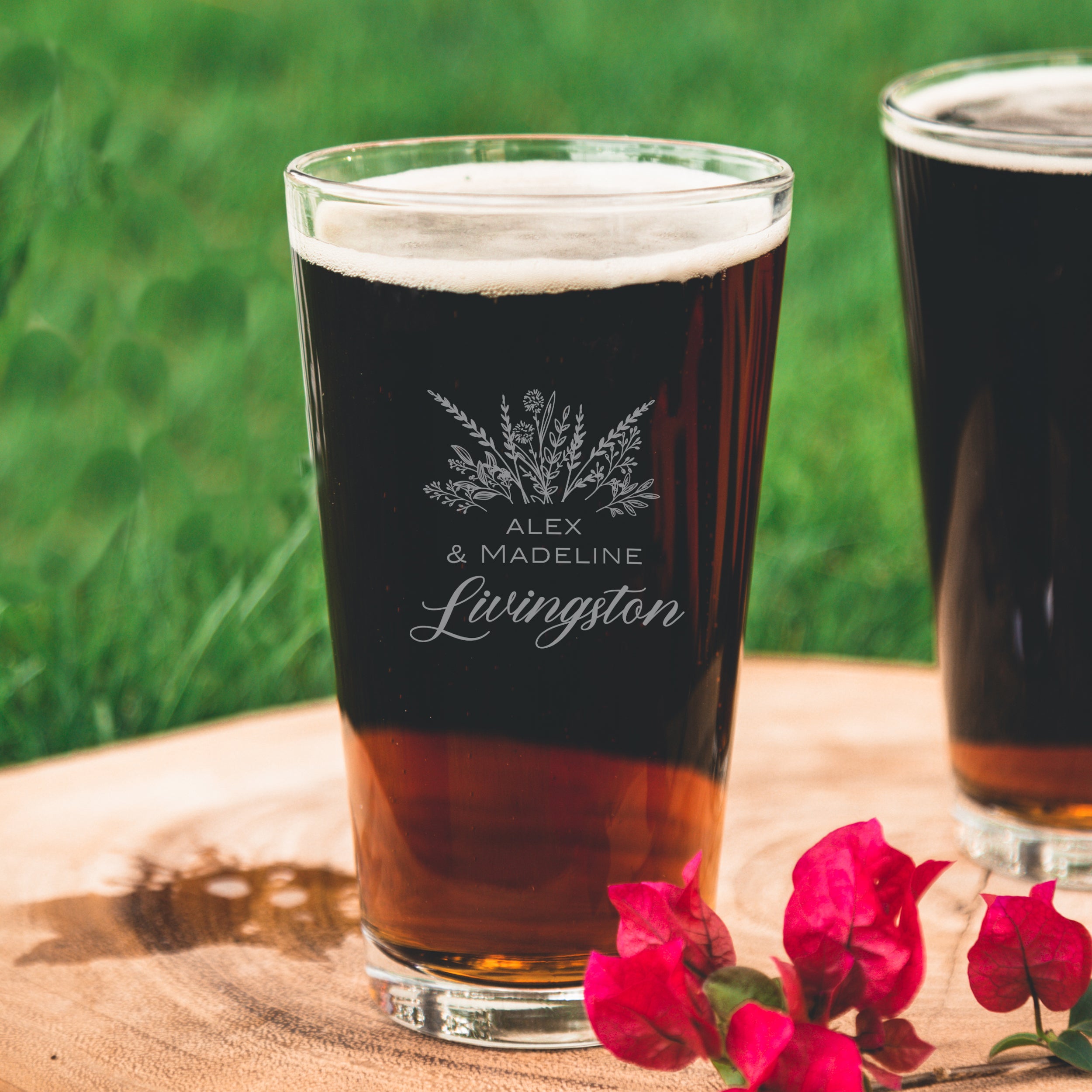 Etched pint glass on a table. The etched design is centered on the glass. The design is of a bouquet of flowers and beneath it is "Alex" and beneath that is "& Madeline" and beneath that is "Livingston". So, three lines of text: first name, send name and a last name.