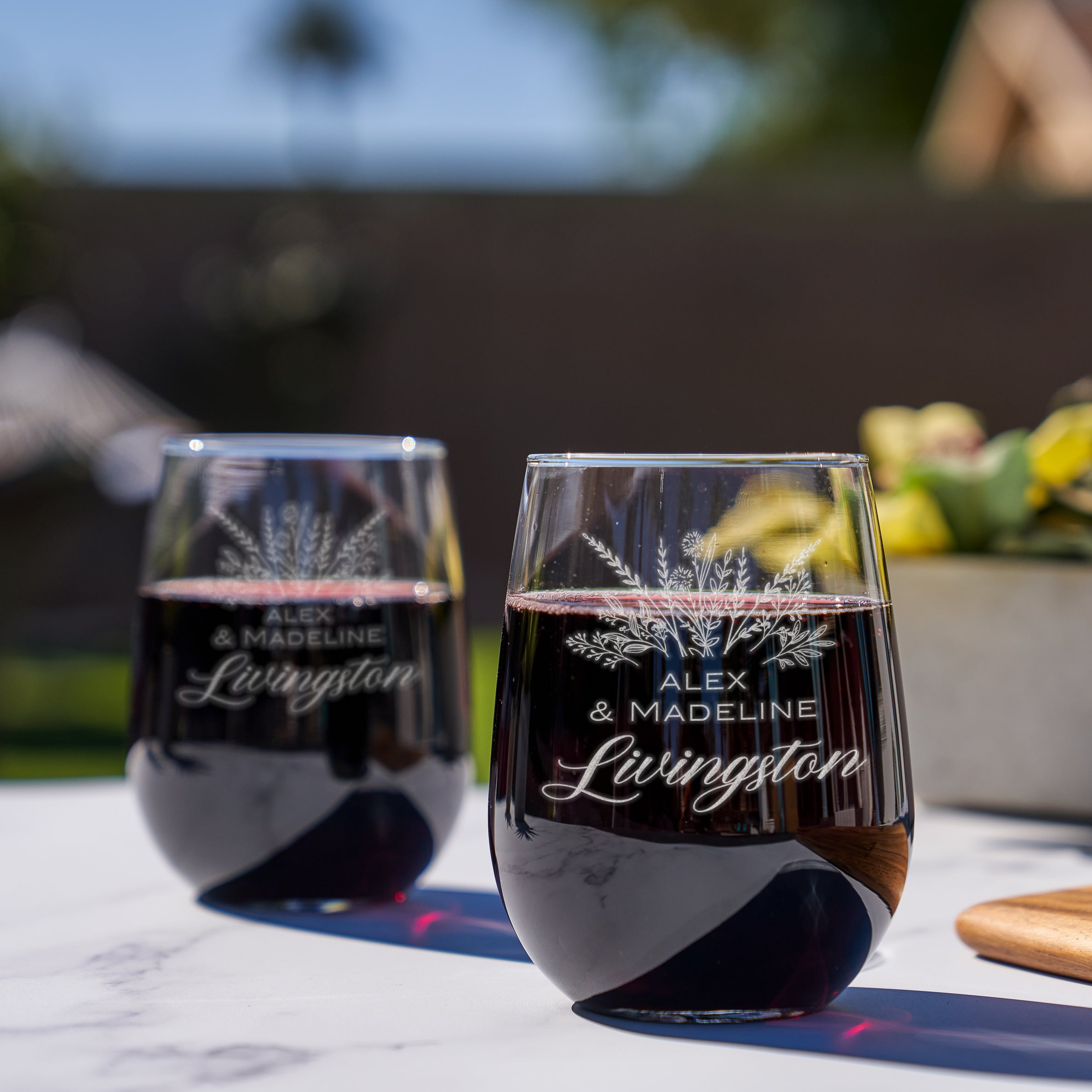 Stemless red wine glass on a table. The etched design is centered on the glass. The design is of a bouquet of flowers and beneath it is "Alex" and beneath that is "& Madeline" and beneath that is "Livingston". So, three lines of text: first name, send name and a last name. The first two names are in print font, the last name is in cursive.