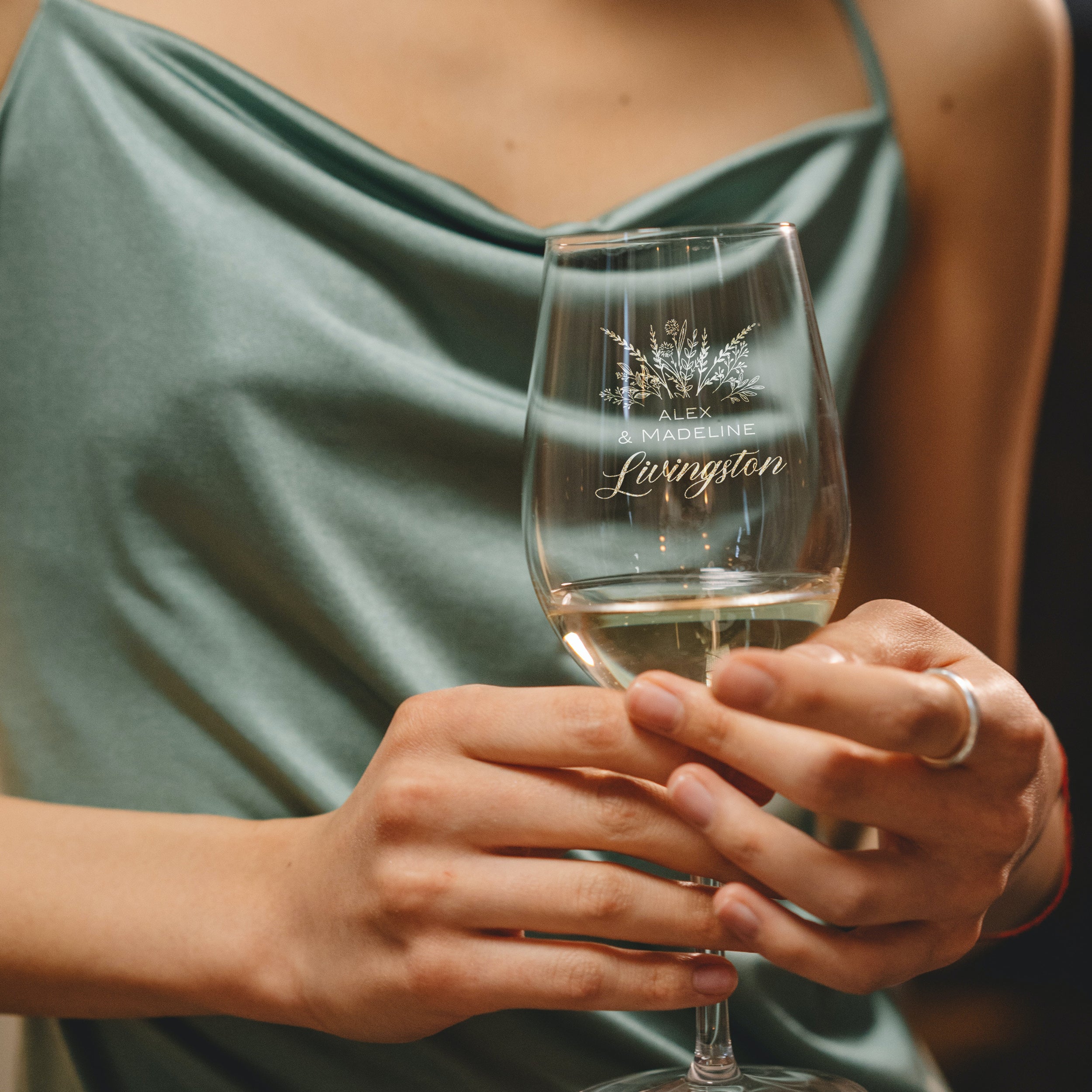 Stemmed white wine glass on a table. The etched design is centered on the glass. The design is of a bouquet of flowers and beneath it is "Alex" and beneath that is "& Madeline" and beneath that is "Livingston". So, three lines of text: first name, send name and a last name. The first two names are in print font, the last name is in cursive.