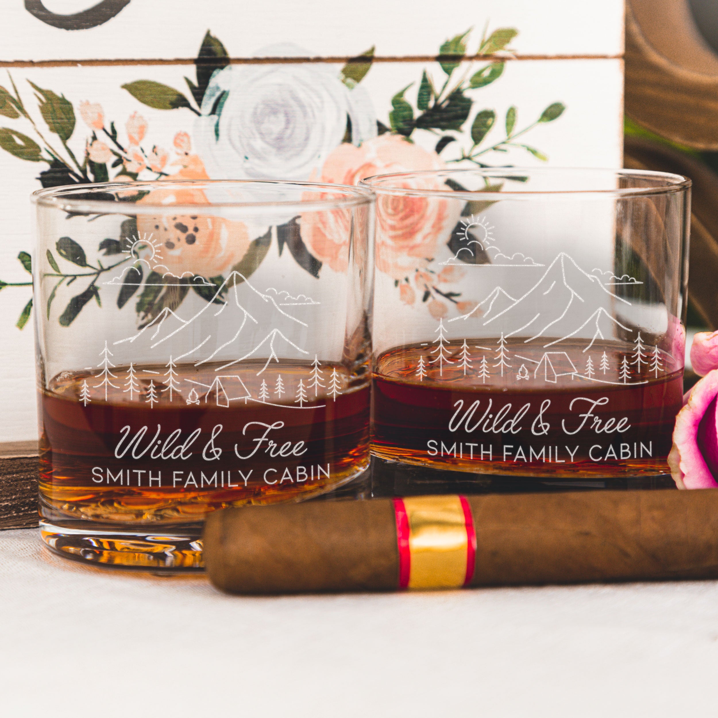 Whiskey glass on a table. The glass has an etched design centered. The design is outdoorsy, of a sun, clouds, mountains, trees and a cabin. Below the outdoors image is "Wild & Free" in cursive and below that is "SMITH FAMILY CABIN" in print font.