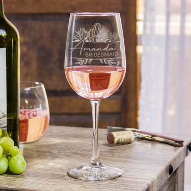 Stemmed wine glass on a table. The glass has a design centered etched in the glass. The design is of different desert cactus plants and below is two lines, and between the lines it says the name "Amanda" in cursive and below the name is "BRIDESMAID" in all caps printed font. 