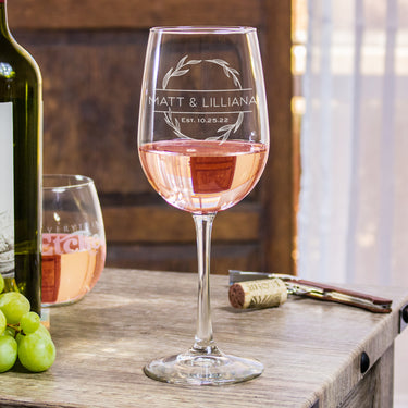 Stemmed wine glass, with an etched design centered. The design is a circle wreath, in the middle is two lines breaking up the wreath and has "MATT & LILLIANA" in printed font all caps. Still inside the wreath, and below the names is "EST. 10.25.22". 